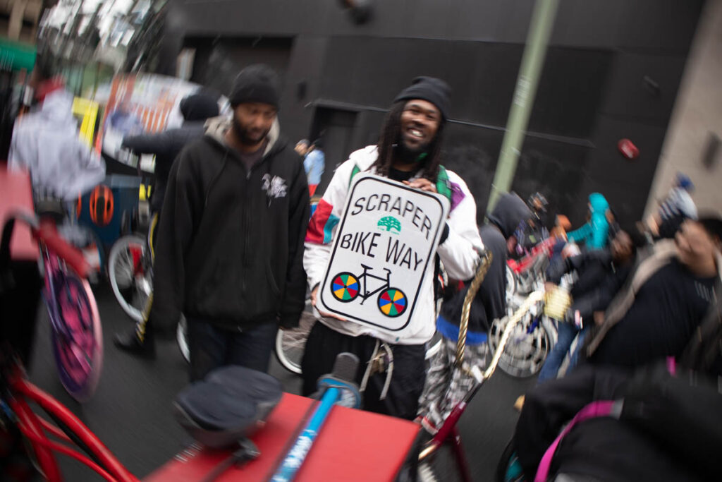 Radially-blurred image of a group of Black cyclists in a city street. In the center of the image, unblurred, one man is holding a road sign which reads "SCRAPER BIKE WAY", with an image of a bicycle with multi-colored wheels, and the City of Oakland logo.