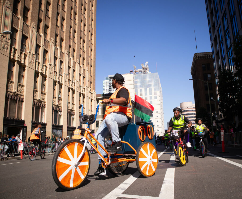 A man on an adult-sized tricycle, with wheels covered decoratively with orange and white tape, rides through downtown Oakland. His trike has a sound system on the back, and a red, black and green flag. He is followed by several children on bikes, apparently riding in a circle.