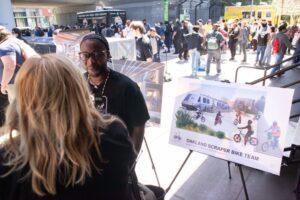 A black man with cornrows, wearing a Scraper Bike Way t-shirt, being interviewed by a blonde woman holding a "KTVU" microphone. He stands in front of a poster board reading "Oakland Scraper Bike Team", with an image of a BART car converted into a community space, with many kids on colorful bikes riding around.