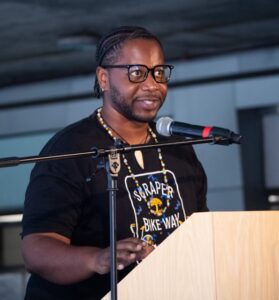 A Black man with a t-shirt reading "Scraper Bike Way" stands at a podium with a microphone.