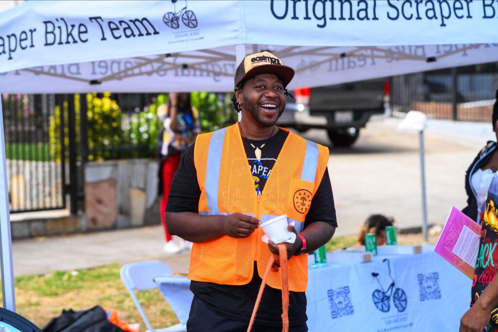 A smiling black man wearing an orange traffic safety vest stands in front of a pop-up canopy reading "Original Scraper Bike Team." It appears to be at a public event; a table is set up under the canopy, which appears to be on a street.