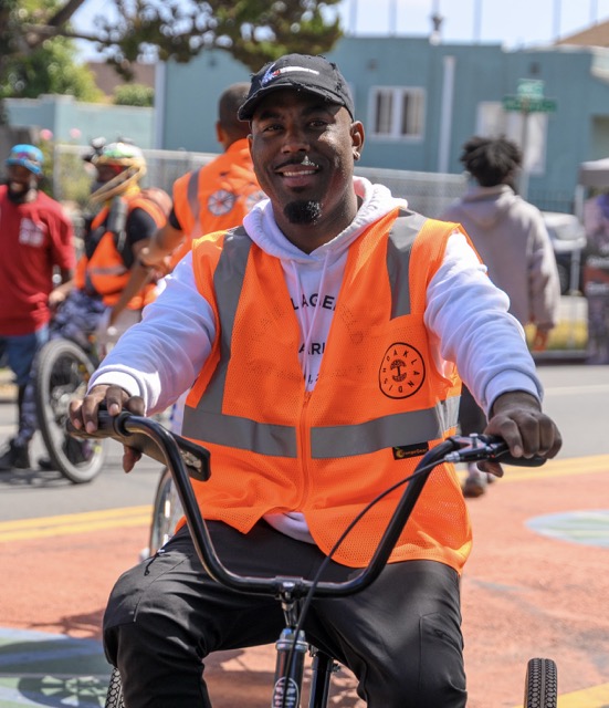 Photo of a smiling Black man wearing an orange safety vest, sitting on a cargo tricycle. In the background are several other people, some with similar orange vests.