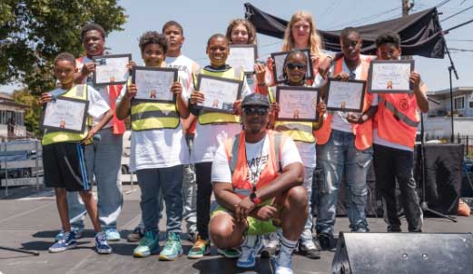 A group of kids wearing orange safety vests smile for a picture while holding up membership certificates. A Black adult man crouches in front of the group.